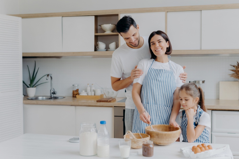 Happy family cook together at kitchen. Father, mother and dauther busy preparing delicious meal at home. Husband embraces wife who whisks and prepares dough, bake cookies. Food, togetherness