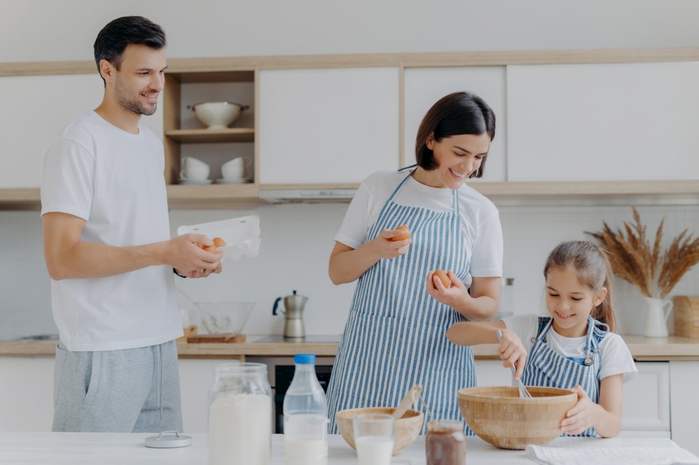 Mother and father give eggs to daughter who prepares dough, busy cooking together during weekend, have happy moods, prepare food. Three family members at home. Parenthood and togetherness concept