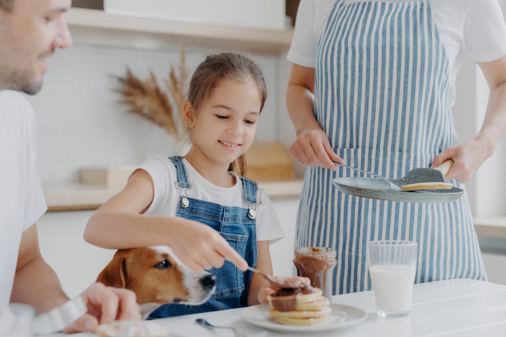 Glad small girl enjoys eating tasty dessert prepared by mum, adds melted chocolate to pancakes, enjoys being together and mother, father and dog, have delicious nutritious breakfast in kitchen