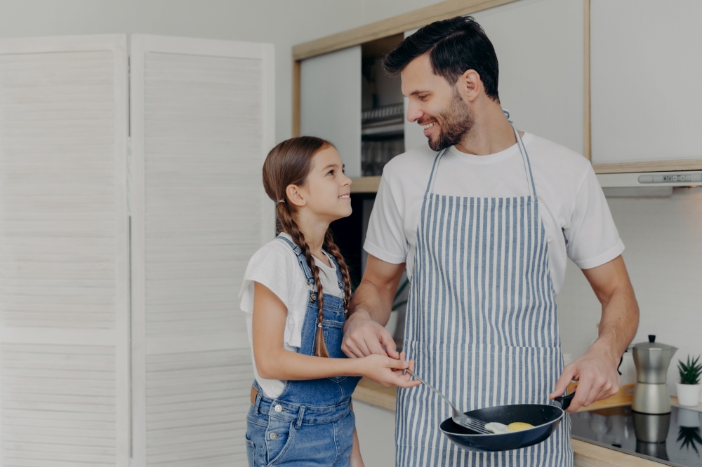 Affectionate father in apton fries eggs, teaches small daughter how to cook, pose together at kitchen, prepare delicious breakfast, look at each other with smile. Children, fatherhood, domestic life