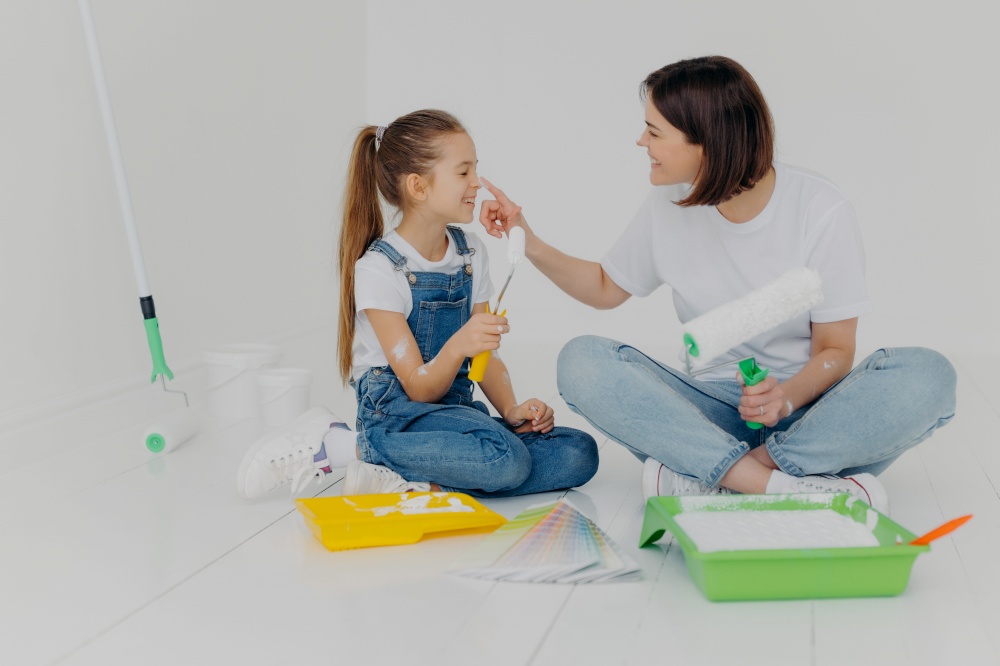 Happy brunette woman plays with daughter, have fun after painting and refurbishment of room, smeark each other with paint, choose color from sample, hold paint rollers, sit on floor in casual wear