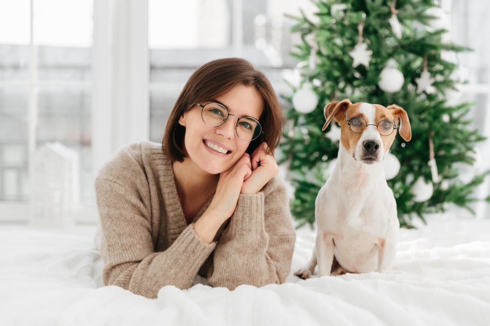 Beautiful brunette adult woman wears big round spectacles, tilts head, wears warm winter sweater, her pet in glasses poses near at bed, express happy emotions, await for Christmas, enjoy coziness