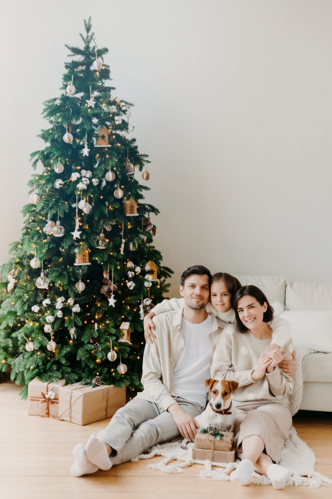 Three family members pose on floor in cozy room, gift boxes around, decorated New Year tree and sofa. Father, mother and daughter with jack russell terrier dog anticipate for coming holiday.