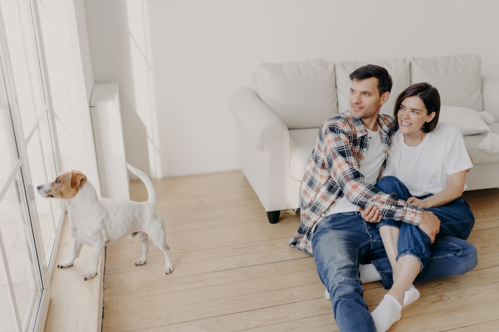 Pleased satisfied romantic couple embrace and looks out of window, pose in empty room near sofa, their dog wants to have outdoor walk. Woman and man enjoy spare time at home, live in new flat