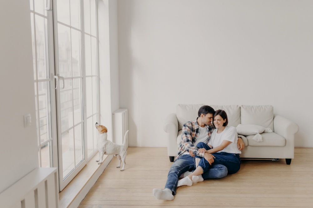Satisfied husband and wife have romantic relationship, sit on floor near white couch in big room, wear jeans, tshirts and socks, spend leisure time in domestic atmosphere, dog poses near balcony