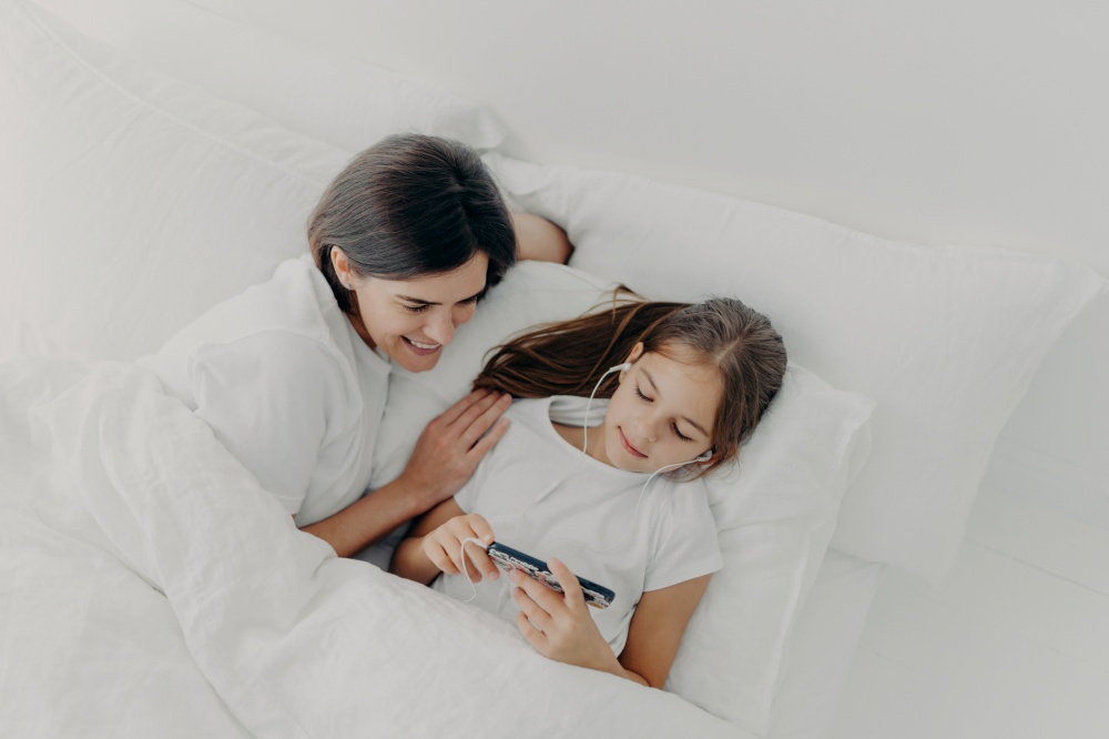 Shot of happy mother spends free time with little lovely daughter who is focused in smartphone, listens song in earphones, pose in bed with white soft bedclothes, have lazy morning during weekend