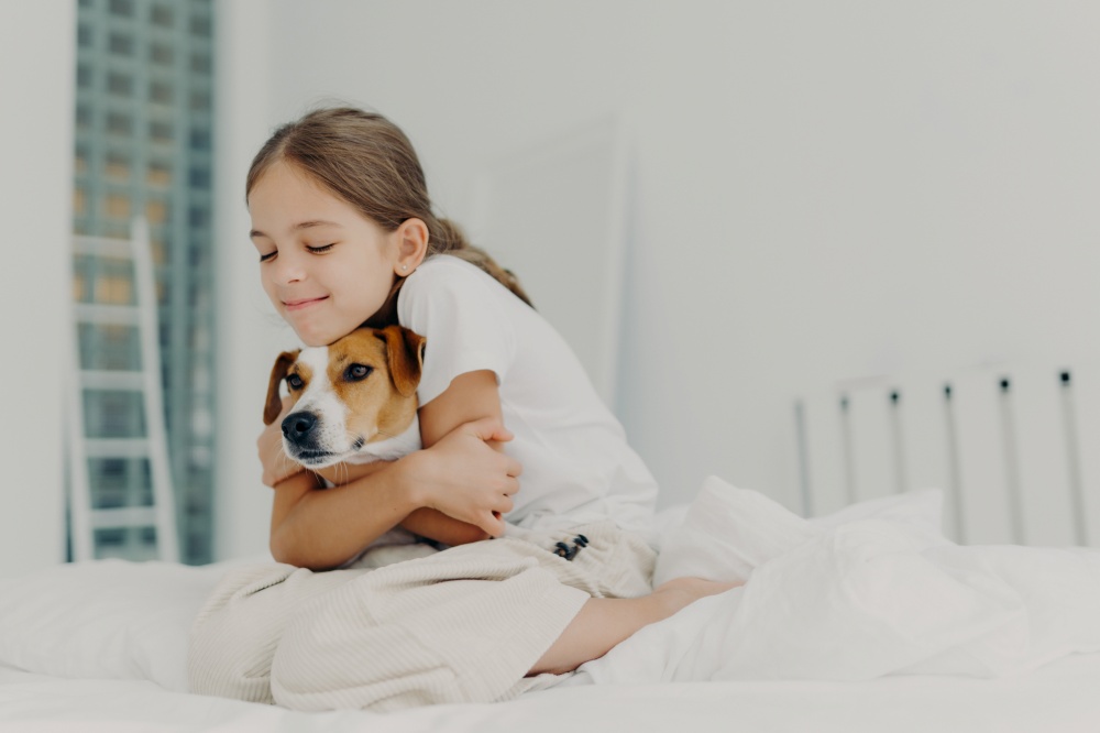 Pleased little girl plays with pet, embraces dog and keeps eyes closed from pleasure, dressed in nightclothes, poses on bed after waking up, expresses love to pedigree animal. Bedding concept