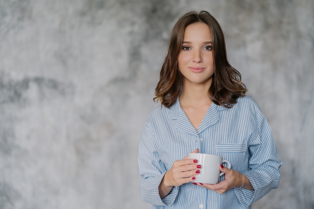 Attractive woman dressed in nightwear, holds white mug with coffee or tea, has morning drink, poses indoor against blurred background with copy space for your advertisement or promotional content