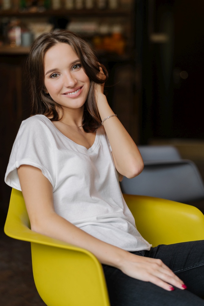 Vertical shot of pretty girl dressed in white t shirt and jeans, has happy look at camera, sits at yellow armchair, being in good mood, feels carefree, has pleasant smile on face. Feminity and rest