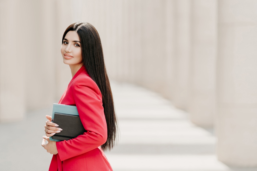 Sideways shot of elegant brunette lady with dark long straight hair, dressed in fashionable red suit, has manicure, carries diary, stands outdoor over white building. Female employee in formal apparel