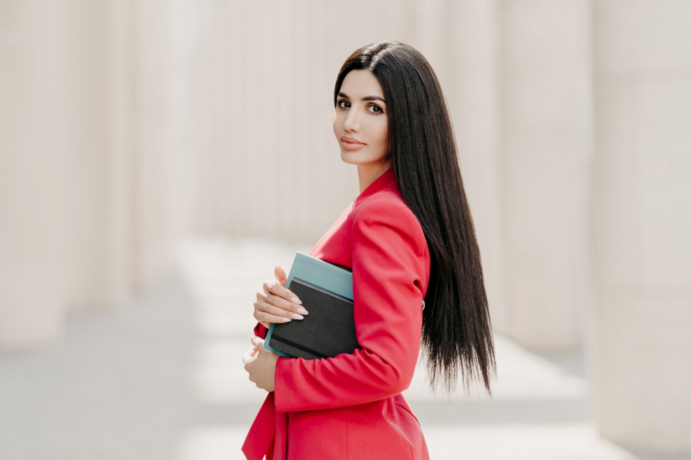 Serious brunette businesswoman with makeup, manicure, wears red jacket, carries diary and notepad, stands in profile outdoor, goes for work. Elegant businesslady on street. Stylish director outside