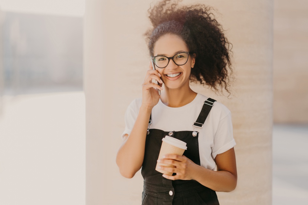 Pleasant looking curly teenage girl wears transparent glasses, talks on mobile phone, has coffee break, dressed in white t shirt and overalls, enjoys spare time and conversation. People and technology