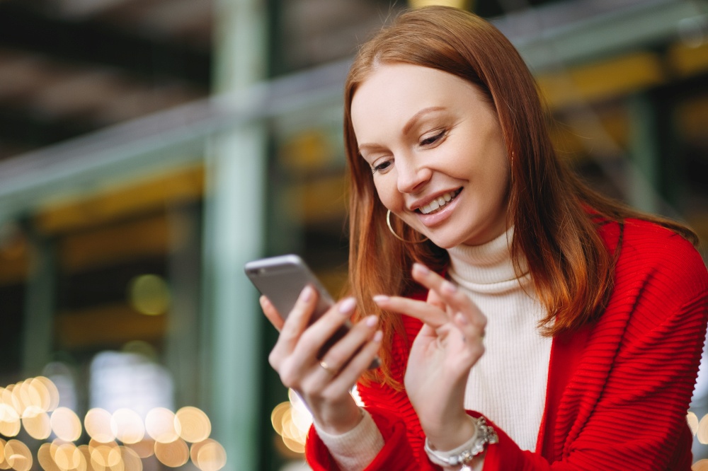 Positive pleased female model uses mobile phone data, uses chat application, holds modern cell phone, smiles positively, dressed in red coat, waits for answer or call from boyfriend, has brown hair