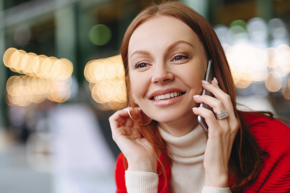 Headshot of attractive young woman with glad facial expression, talks via cell phone, has brown hair, happy to hear news, has free time, blurred background with copy space for your promotion