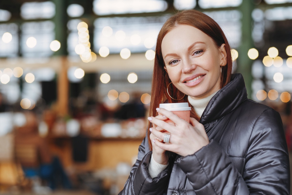 Horizontal shot of lovely woman with toothy smile, holds paper cup of drink, enjoys aromatic coffee, wears fashionable jacket, poses over blurred background with copy space. Street style concept