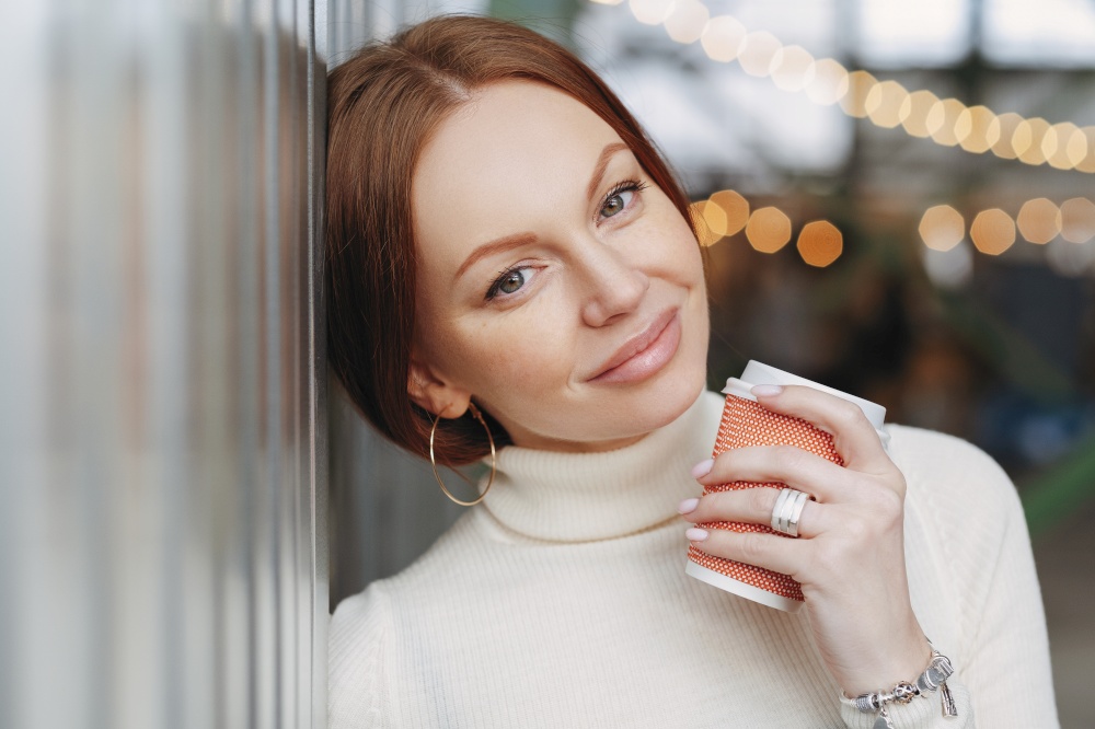 Dreamy pensive Caucasian woman with make up, dressed in casual outfit, leans on wall, holds takeaway coffee, remembers pleasant moments during outdoor stroll, has appealing look. Drink concept