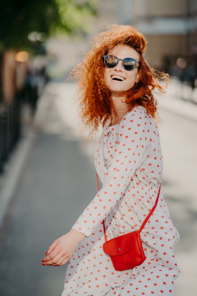 Happy carefree redhead woman laughs positively, spends free time outdoor in city, wears sunglasses, long sleeved dress, carries red bag, has fun, enjoys recreation time. People and lifestyle