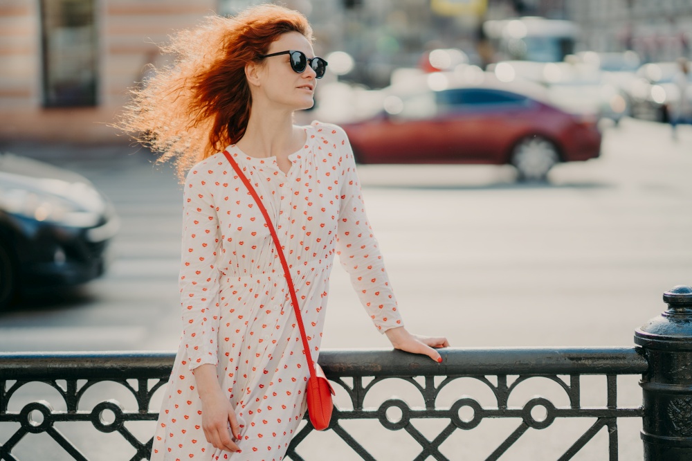 Horizontal shot of red haired woman wears sunglasses, focused aside, poses near hence at street, poses against road with transport, strolls in city during summer sunny day, copy space for your text