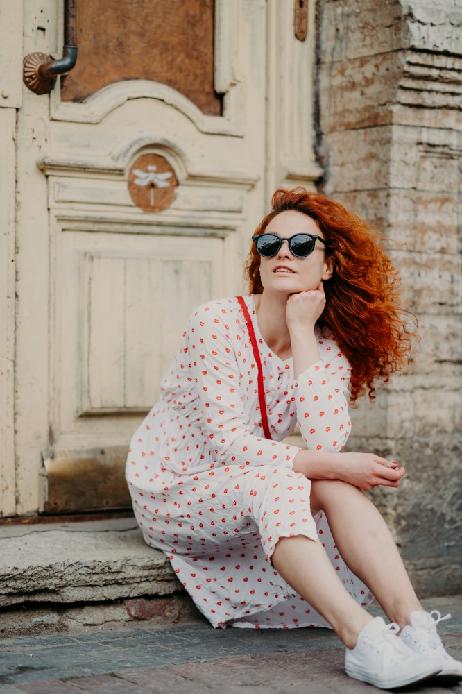 Red haired thoughtful woman has rest after strolling, poses near old building, sits on threshold, wears sunglasses, dress and white sneakers, explores ancient places in European city. Vertical shot