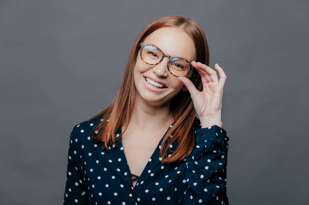 Smiling woman with pleased facial expression, keeps hand on rim of spectacles, wears black polka dot blouse, rejoices promotion at work, poses over grey background. People, happiness concept
