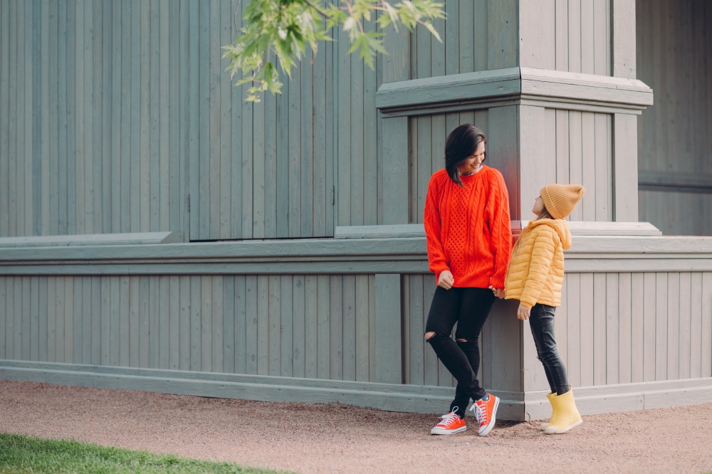 Outdoor shot of friendly woman holds daughters hand, stand at corner of some building, wear warm clothes, enjoy good day and walk in open air, have recreation time. Happy mum and female kid.