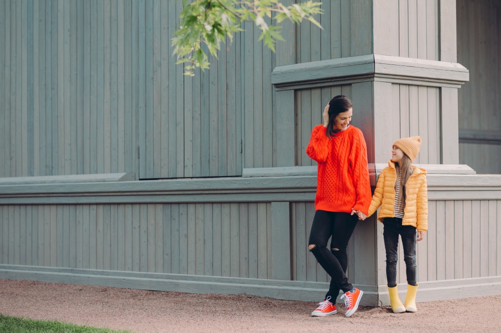 Horizontal view of fashionable European female in red loose sweater, ripped jeans, holds hands together with her younger sister, pose outside together, have walk in open air, have friendly relations