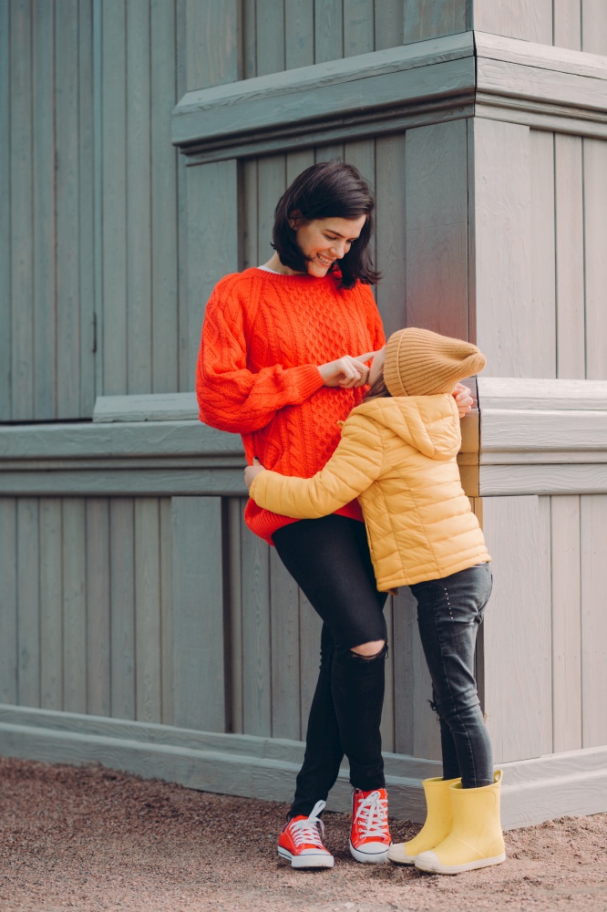 Photo of happy young mum touches nose of her curious pleased daughter, stand closely to each other at corner of wooden building. Lovely small child wears hat, jacket and boots, has fun with mother