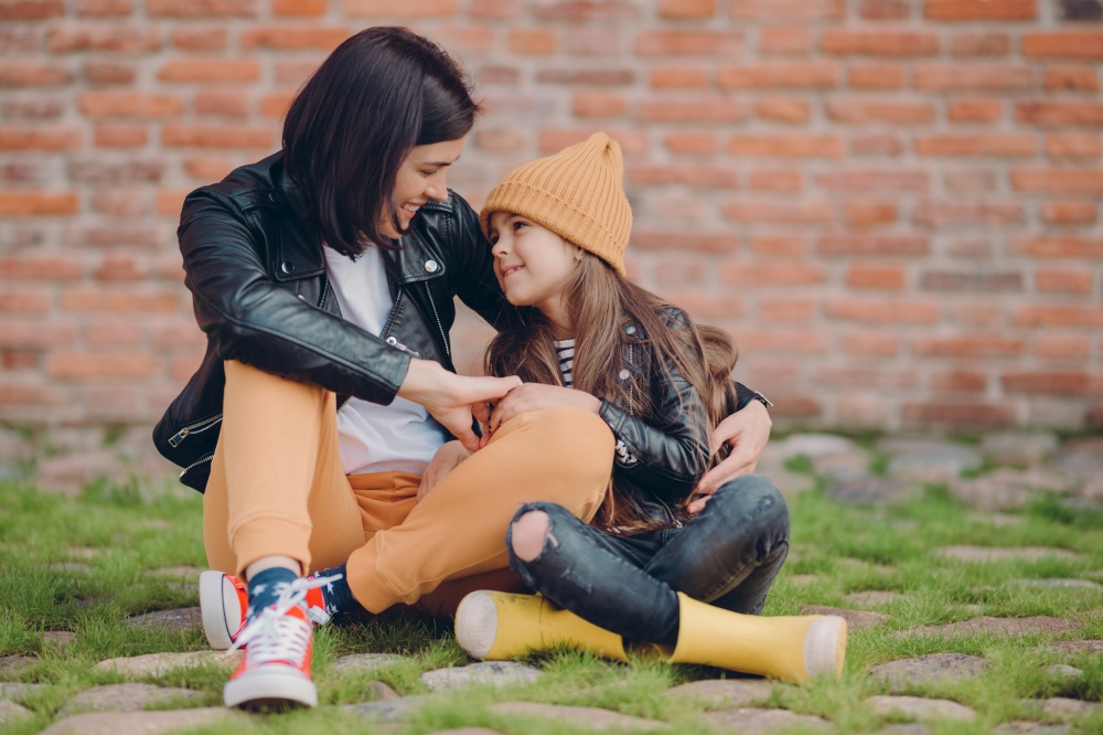Lovely family of young mother and her small daughter embrace friendly, have good relationships, dressed in fashionable clothes, pose against brick wall outdoor. Parenthood and childhood concept