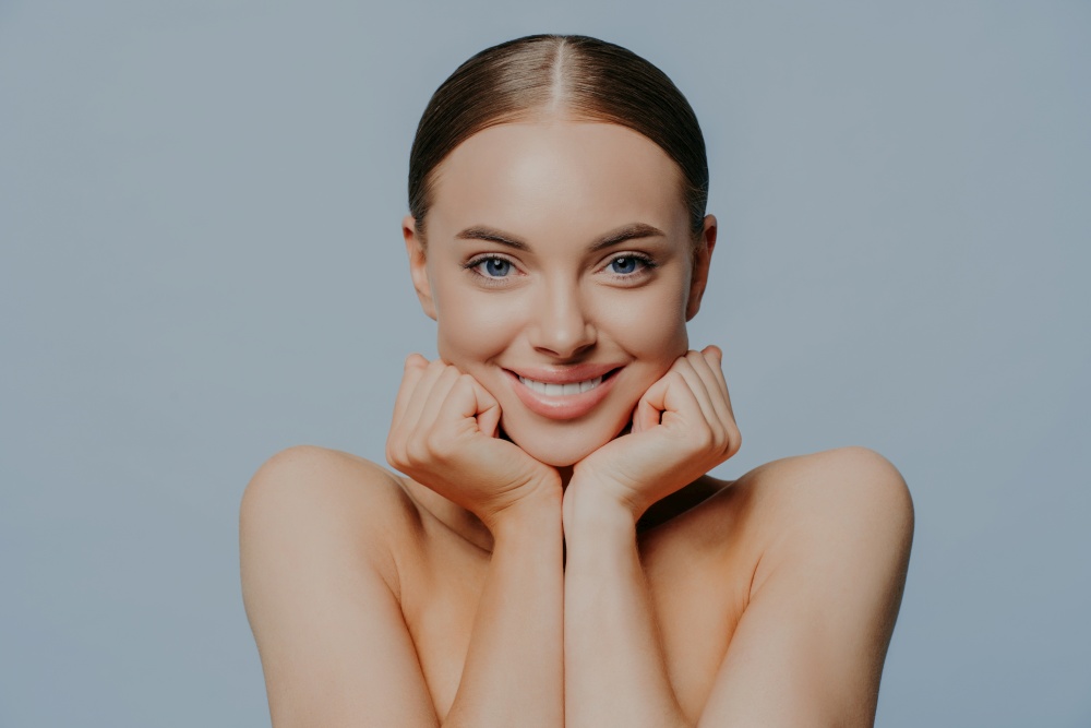 Women, skin care and tenderness concept. Young female takes care of skin and body health, keeps hands under chin and smiles gently, wears makeup, has combed hair, delighted with skin care product