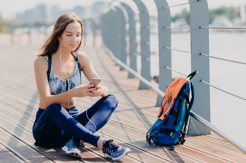 Horizontal shot of beautiful happy atthletic female in sneakers, sportswear, feels relaxed while sits outdoor, uses modern cell phone for surfing internet, dowloads music for playlist, listens song