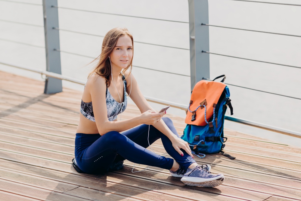 Relaxed lovely woman with dark hair, healthy pure skin, dressed in sportswear, holds cell phone for communication, listens favourite track, rucksack stands near her. People, sport, recreation concept