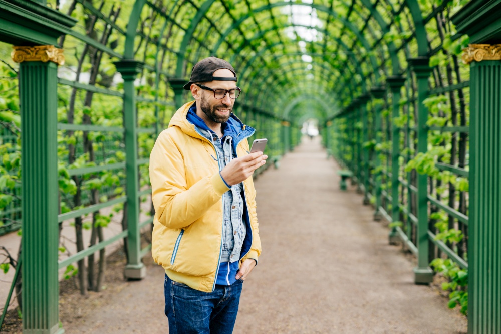 Trendy guy with beard and moustache wearing anorak, jeans and cap standing sideways having attentive look into his smartphone isolated over green arch. Human, style and modern technology concept