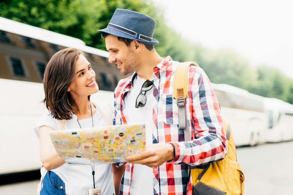 Attractive male and female tourists, being in unfamiliar place, holding map or city guide, deciding where to go first, looking happily at each other, standing near tourists` bus. Travelling concept