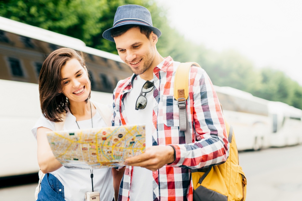 Young family tourists looking attentively in map while searching for museum in big city. Stylish man in hat and checkered shirt holding rucksack, standing near his wife, looking for new destination