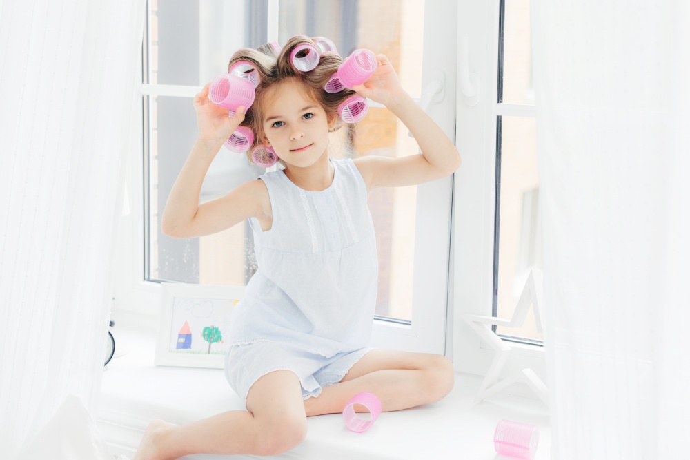Photo of adorable cute girl with curlers on hair, sits on window sill at home, going to have curly hairstyle, wears casual clothes, looks directly into camera. Children, leisure and beauty concept