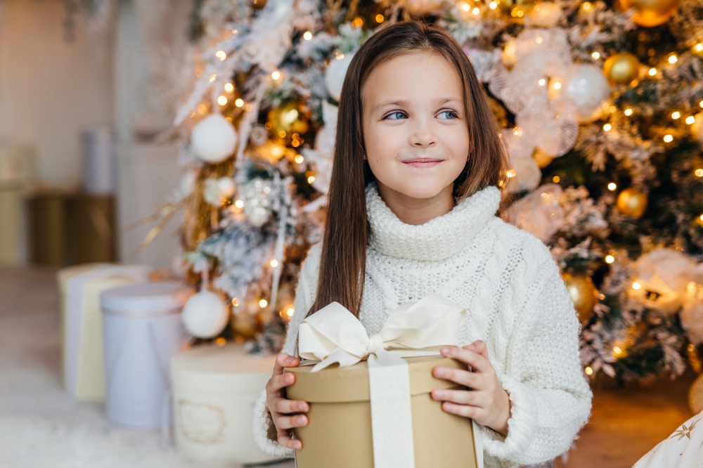 Cute lovely small kid with charming appearance, glad to recieve Christmas gift, looks aside with happy expression, has intrgue what present is, poses near beautiful decorated fir tree.