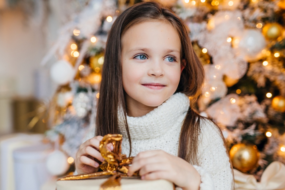 Adorable little female child with warm blue eyes, long dark hair, wears knitted warm white sweater, holds present stands against decorated New Year tree, looks aside on her affectionate parents