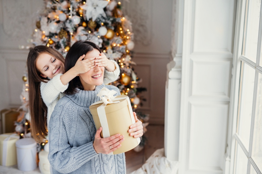 Portrait of little girl closes mother`s eyes, congratulates her with New Year or Christmas, stand near window in living room, have real miracle and feeling of holiday. Winter, celebration, season