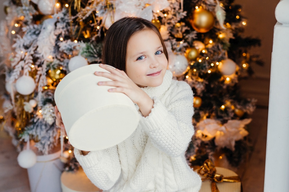 Blue eyed pretty lovely small child holds present box, wonders what is inside, stands near New Year or Christmas tree, recieves surprise from parents. Delighful kid with gift. Children and holidays