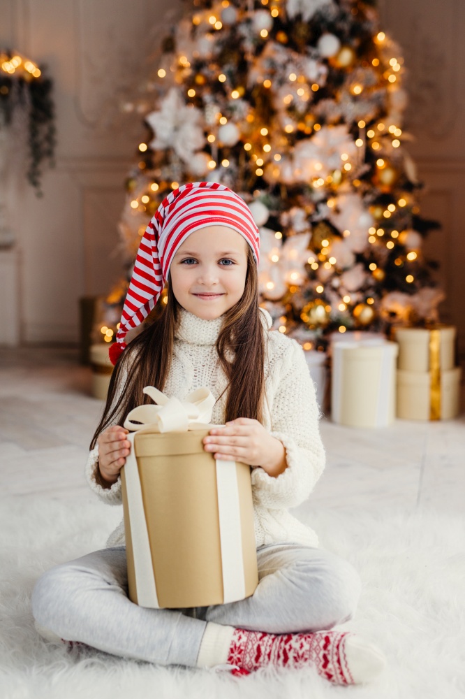 Vertical portrait of beautiful pleasant looking small child wears knitted sweater and socks, sits crossed legs with present, has desire to wrap it, being in living room near decorated New Year tree