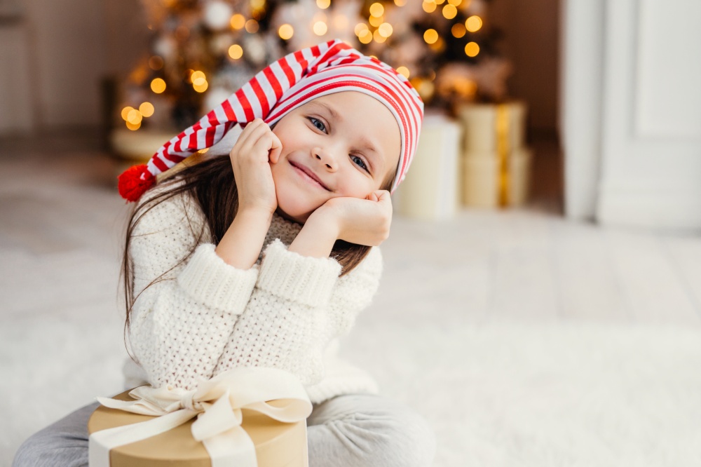 Beautiful small child poses in living room, leans at present gift, has happy expression, glad to recieve surprise from parents, spends holidays in family circle. Merry Christmas and happy New Year
