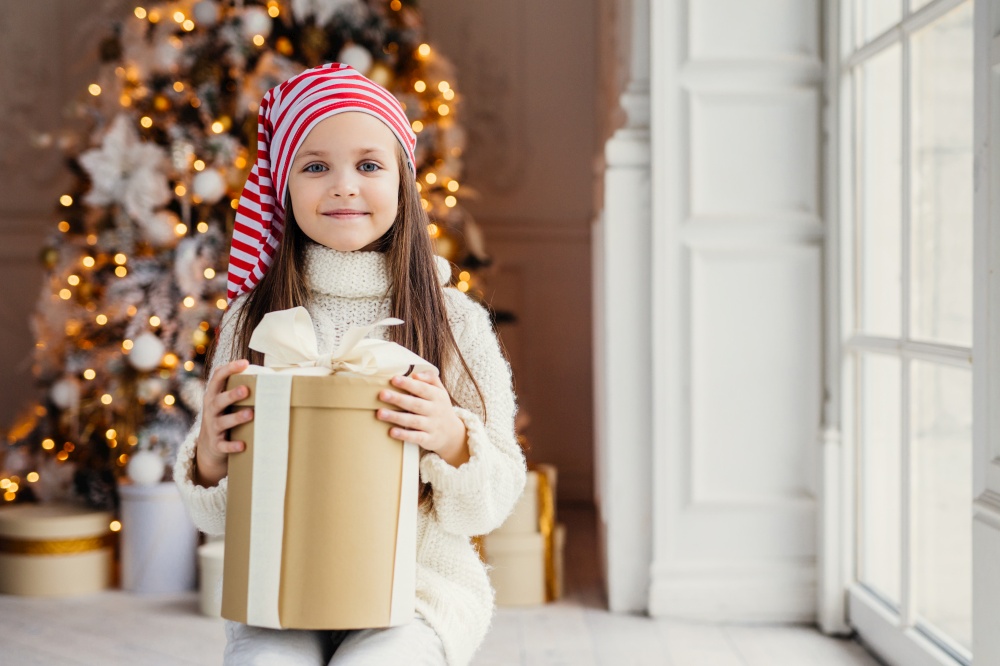 Indoor shot of pleasant looking small kid with blue charming eyes, wears santa hat, holds present in wrapped box, sits over decorated New Year background. Childhood, celebration concept