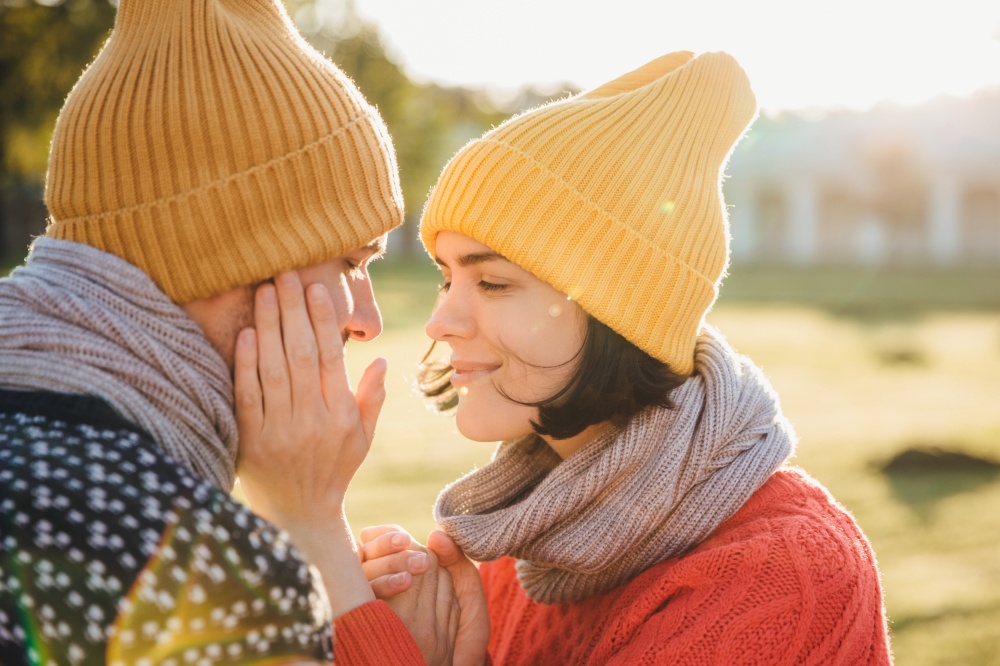 Horizontal portrait of brunette female in warm hat, scarf and sweater, keeps hands on boyfriend`s cheek, have romantic moment, going to kiss as dating. Couple in love look at each other passionately