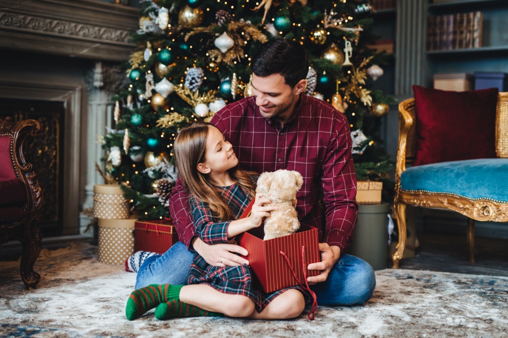 Small female child holds teddy bear, recieves unexpected gift from affectionate father, being thankful to him. Young dad gives present to daughter, congratulates her with Christmas or New Year
