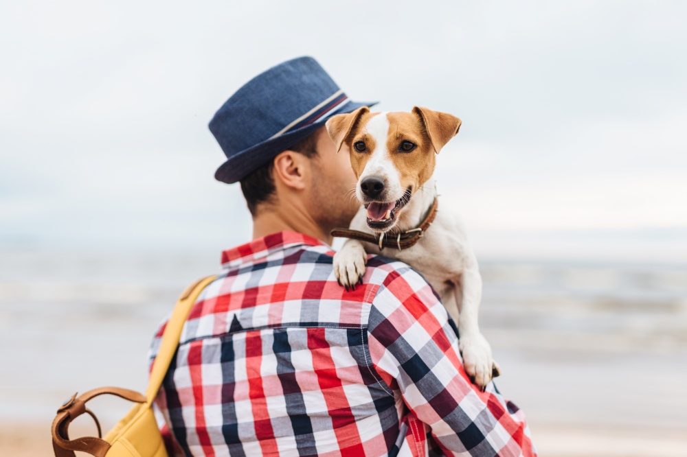 Small beautiful dog on owner`s hands. Stylish young male model dressed in fashionable clothes carries his pet and bag, stand at seashore, come to admire wonderful landscapes and breath fresh air