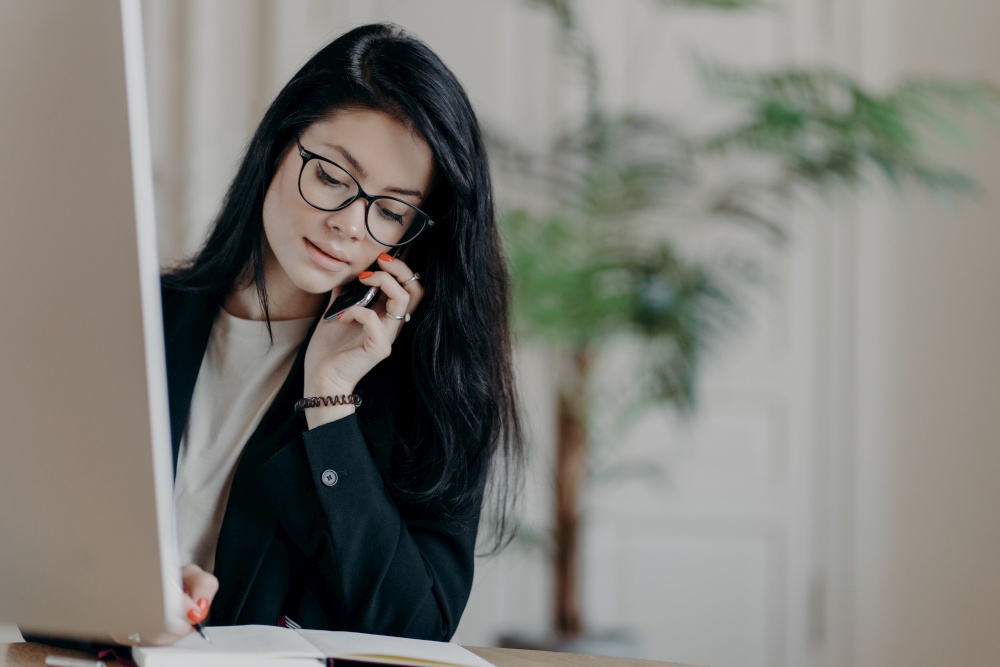 Stylish female entrepreneur in black suit, has phone conversation, makes notes in organizer looks assured and ambitious poses at desktop against office interior. Young woman manager talks via cellular