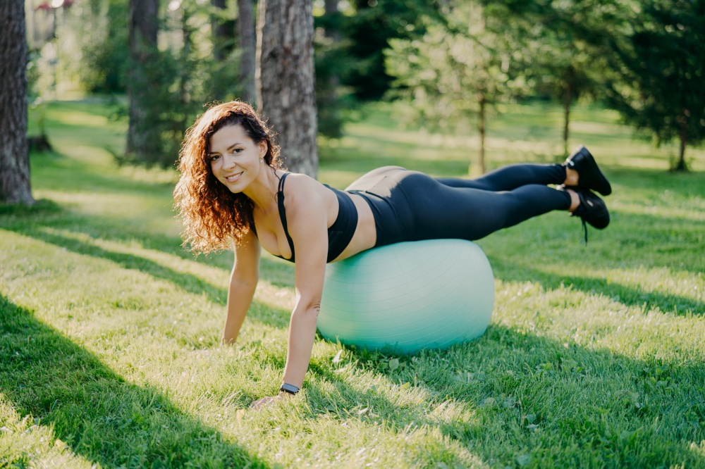 Sport, recreation, fitness and healthy lifestyle concept. Brunette sporty young European woman in active wear leans on fitness ball, makes physical exercises outdoor, enjoys favorite activity