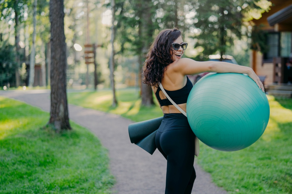 Outdoor shot of happy brunette woman carries fitball and fitness mat, wears sunglasses and sport clothes, poses in green park, being in good physical shape. Healthy lifestyle and hobby concept