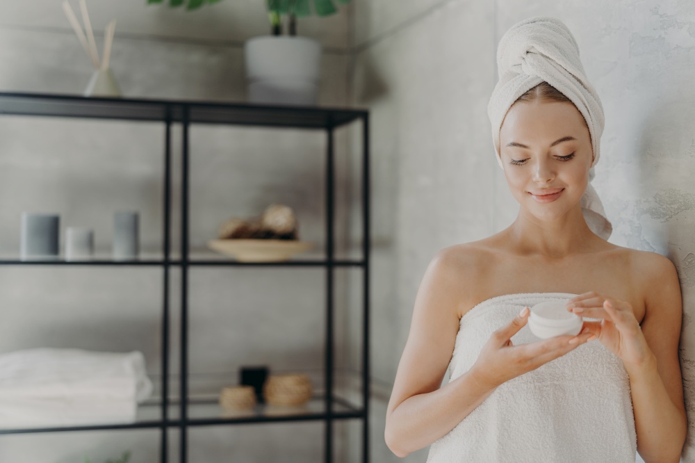 Horizontal shot of pleased woman with healthy skin applies body cream, uses cosmetic product for caring of herself, poses in bathroom after spa bath, undergoes hygiene procedure. Cosmetology concept