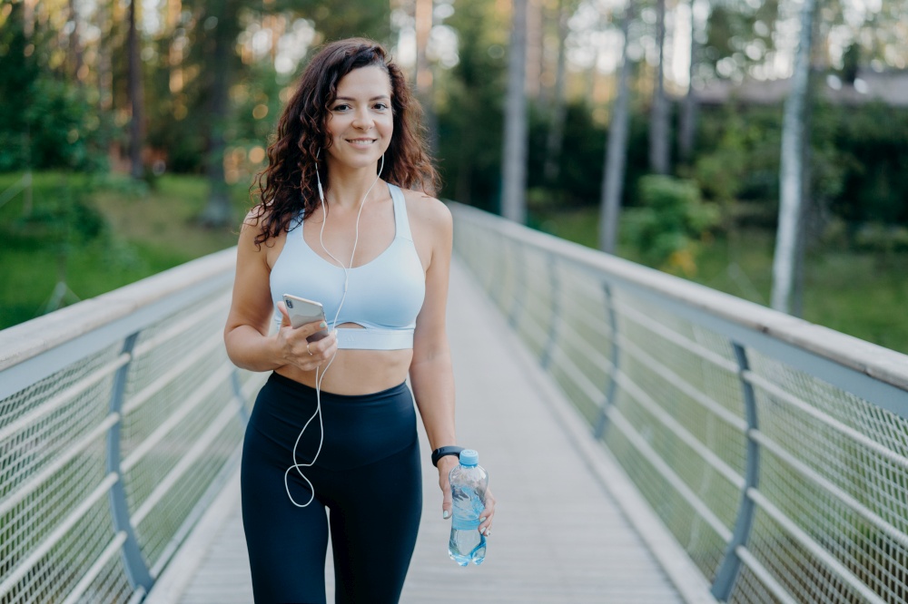 Active beautiful sporty woman in active wear holds mobile phone, listens music in earphones, takes break after jogging holds bottle of fresh water poses at bridge outdoors. Fitness, lifestyle concept